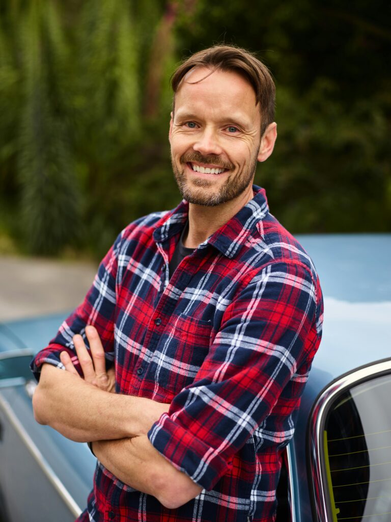 Happy man in plaid shirt with arms crossed, smiling confidently next to a car outdoors.